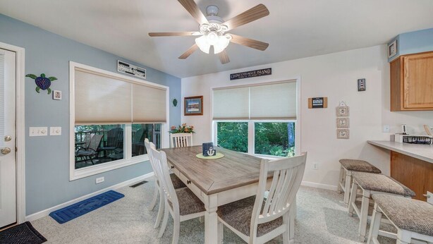 A dining area with a wooden table, white chairs, and a ceiling fan. There are large windows with blinds, a kitchen area with wooden cabinets, and a blue rug on the floor. - Sundance by Seaside Vacations & Sales (Chincoteague Island)
