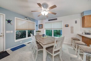 A dining area with a wooden table, white chairs, and a ceiling fan. There are large windows with blinds, a kitchen area with wooden cabinets, and a blue rug on the floor. - Sundance by Seaside Vacations & Sales (Chincoteague Island)