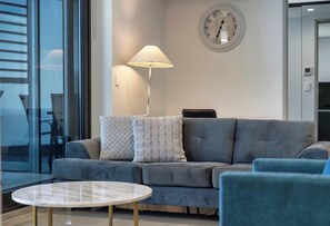 A modern living room with a grey sofa, patterned cushions, a round table, a floor lamp, and a wall clock.