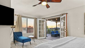 A bedroom with a bed, a blue chair, a small table, and an open sliding door leading to an outdoor seating area. - Elegant Hillside Estate | Heated Pool+Sunset Views (El Cajon)