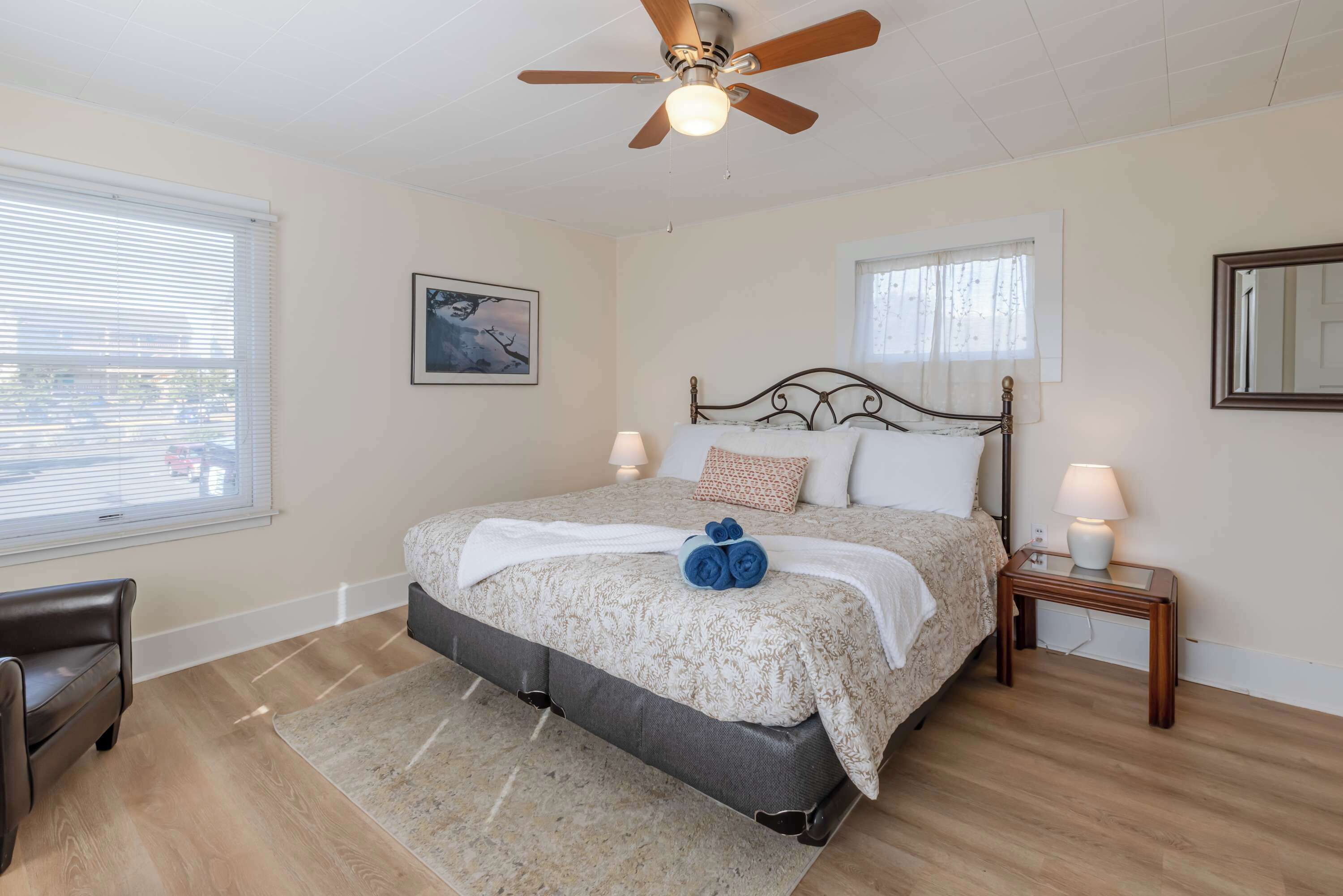 A bedroom with a bed, a ceiling fan, a nightstand with a lamp, a window with blinds, and a chair.