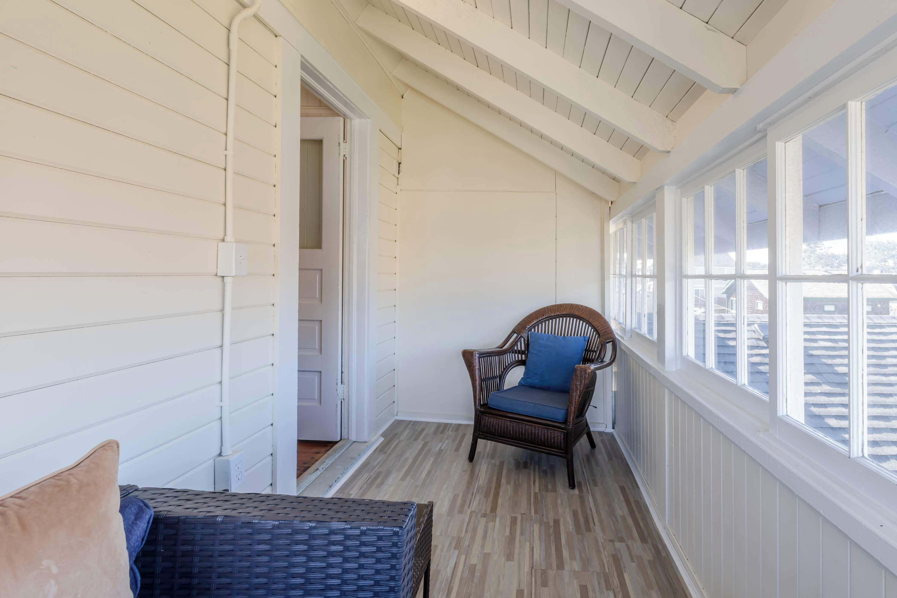 A covered porch with a wicker chair and cushions, wooden flooring, and large windows.