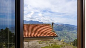 Une vue à travers la fenêtre d’un bâtiment dont le toit est recouvert de tuiles rouges, avec une cheminée et un paysage montagneux en arrière-plan.