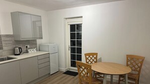 A compact kitchen with a refrigerator, kettle, and cabinets. There is a dining table with two chairs and a window with a door.
