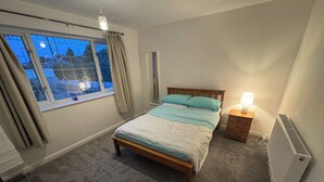 A bedroom with a bed, a nightstand, a radiator, a window with curtains, and a view of the outside.