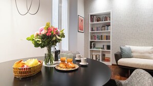 Un coin repas avec une table mise pour le petit-déjeuner, comprenant des croissants, des fruits et du jus. Un vase rempli de fleurs est posé sur la table, et une bibliothèque se trouve à l’arrière-plan.