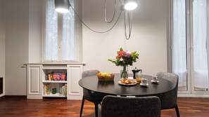A dining area with a round table, chairs, a vase with flowers, a basket of fruit, a teapot, and a pitcher of orange juice. - Italianway Comfort - Vincenzo Monti 29 D (Milan)
