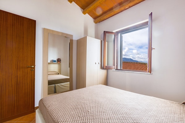 A bedroom with a bed, a wooden wardrobe, a mirror, and a window with a view of a tiled roof.