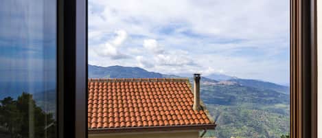 A view through a window of a building with a red-tiled roof, a chimney, and a mountainous landscape in the background.