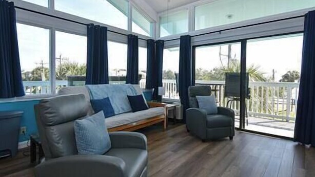 Une pièce spacieuse comprenant un canapé, un fauteuil et une petite table. De grandes fenêtres offrent une vue sur l’extérieur.