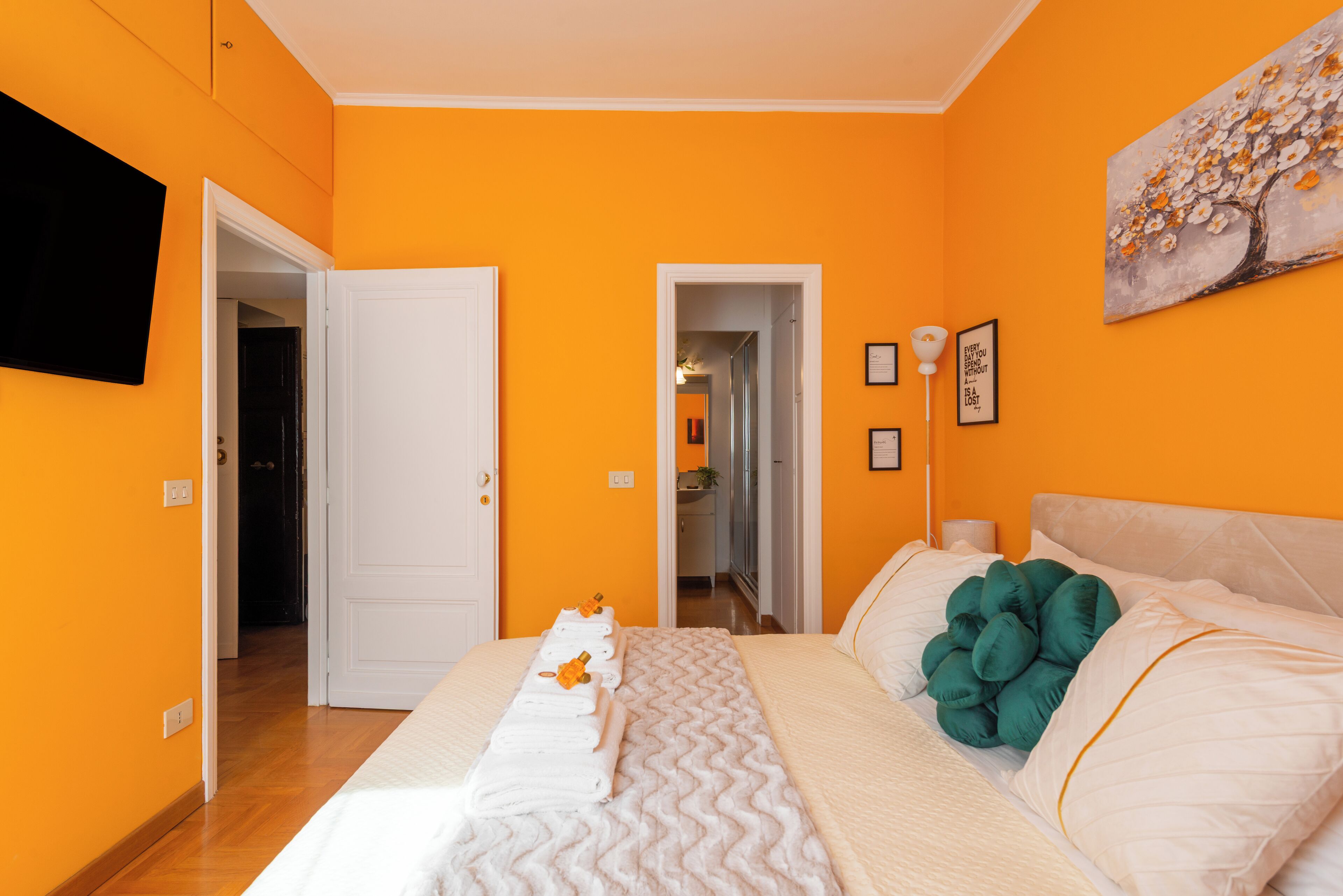 A hotel room with orange walls, a bed with white bedding, a painting on the wall, and a hallway leading to other rooms.
