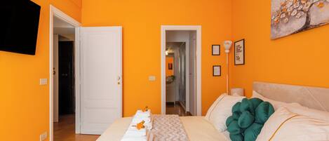 A hotel room with orange walls, a bed with white bedding, a painting on the wall, and a hallway leading to other rooms.