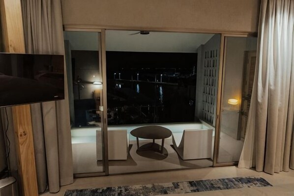 Een kamer met een schuifglasdeur naar een balkon, een flatscreen televisie en een kleine ronde tafel met stoelen.