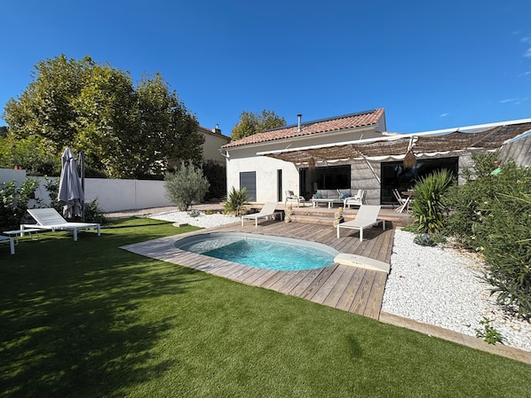 Un jardin avec une piscine, une terrasse dotée de chaises longues et un espace extérieur aménagé et abrité.