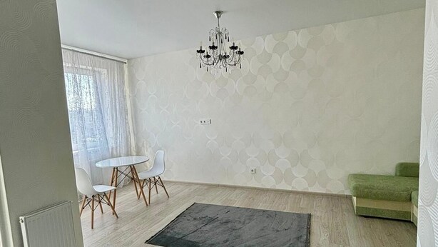 Ein geräumiges Wohnzimmer mit einem modernen Kronleuchter, einem Essbereich mit rundem Tisch und Stühlen sowie einem grünen Sofa.