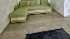 Ein Zimmer mit einem grünen, modularen Sofa, strukturiertem Wandpapier und einem dunklen Teppich auf Holzdielen.