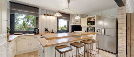 A modern kitchen with a large island, bar stools, a refrigerator, and a view of the outdoors.