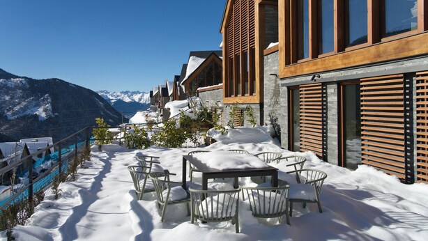 A snow-covered outdoor seating area with a table and chairs, overlooking a mountainous landscape.