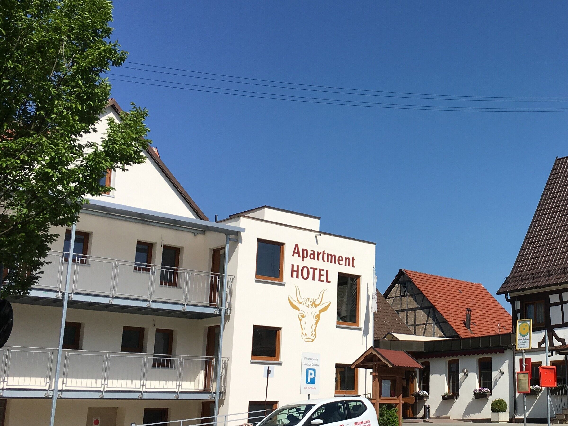 Ein Gebäude mit dem Schriftzug 'Apartment Hotel', einem Balkon, einem Parkschild und einem traditionellen Gebäude mit rotem Dach im Hintergrund.