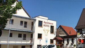 Un immeuble portant l’inscription « Appartement-hôtel », doté d’un balcon, d’un panneau indiquant un parking, et un bâtiment traditionnel avec un toit rouge en arrière-plan.