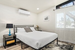 Ein Schlafzimmer mit einem Bett, einem Nachttisch, einer Lampe, einem Fenster mit Vorhängen und einer Klimaanlage.