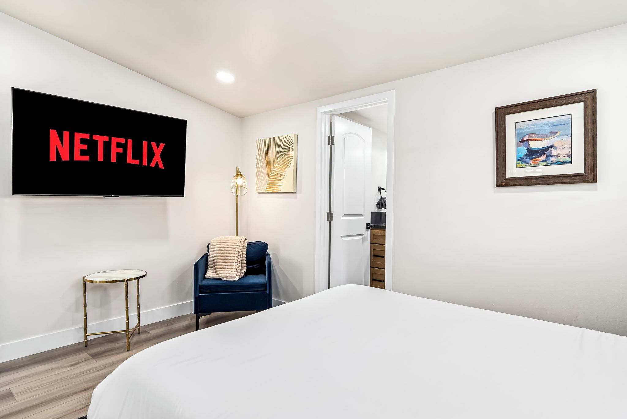 A hotel room with a bed, a TV displaying 'NETFLIX', a small table, a chair, a lamp, and a door.