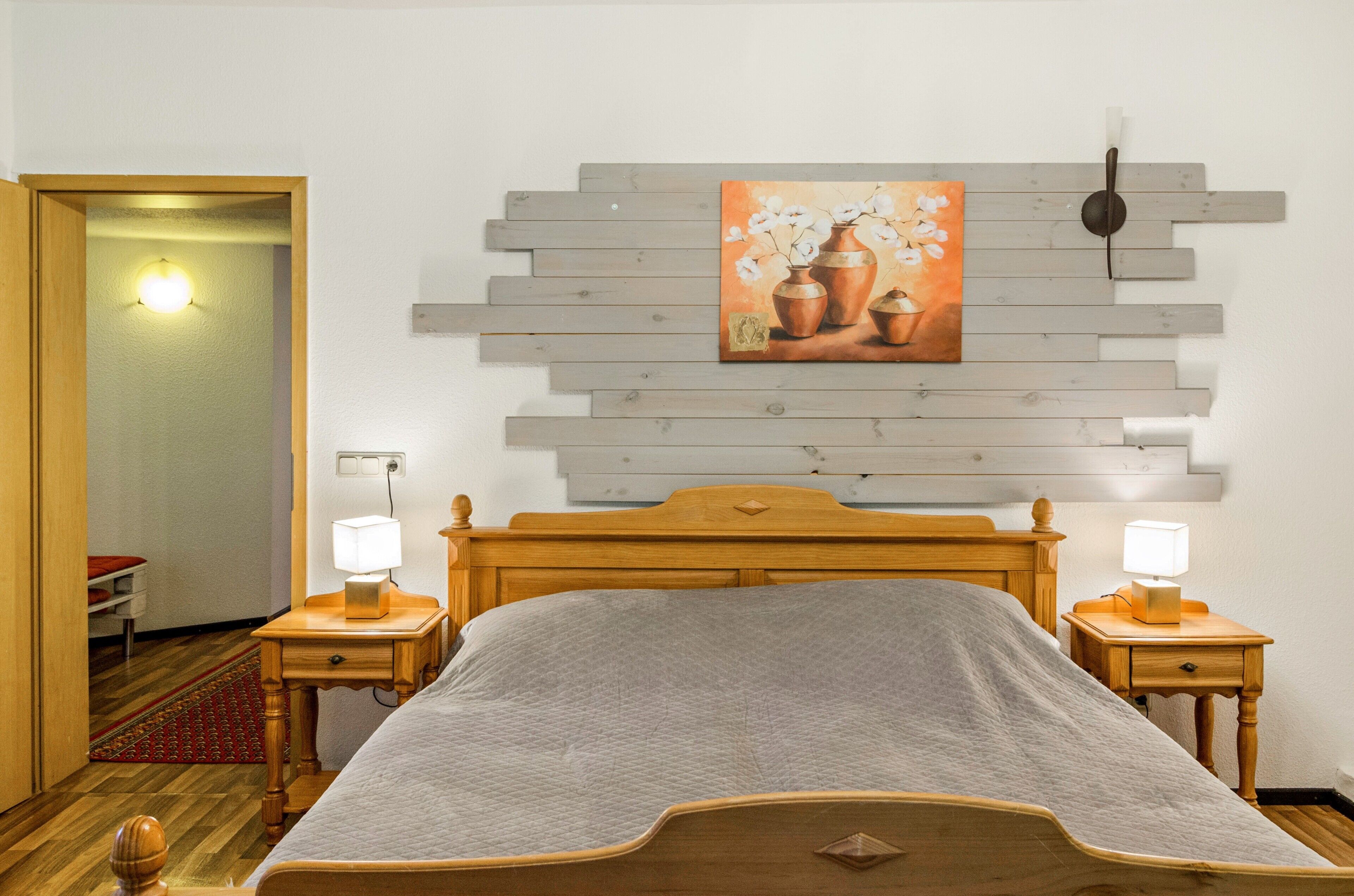 A bedroom with a wooden bed, two bedside tables with lamps, a wall-mounted artwork, and a door leading to another room.