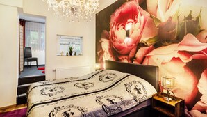 A bedroom with a large floral mural, a chandelier, a bed with a patterned bedspread, a nightstand with a lamp, and a window with a view of the outdoors. - Apartment "Double Room with Private Bathroom" (Stolberg i. Harz)