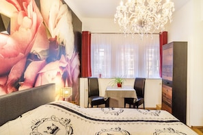 A bedroom with a large bed, a chandelier, a table with chairs, and a window with curtains.
