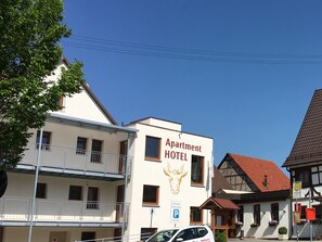Ein Gebäude mit dem Schriftzug 'Apartment Hotel', einem Balkon, einem Parkschild und einem traditionellen Gebäude mit rotem Dach im Hintergrund.