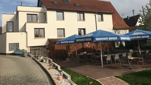 A building with a modern design, featuring a flat roof and multiple windows. There are outdoor tables and chairs arranged for dining, and a blue canopy with the Schneider Weisse logo is visible. - Apartment '6' with Shared Terrace and Wi-Fi (Mössingen)