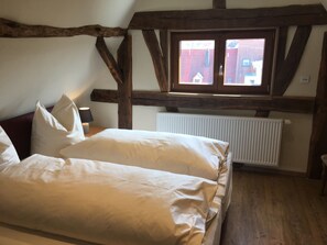 A bedroom with a wooden ceiling, a window with a view of buildings, a radiator, and a bed with pillows.