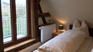 Une chambre avec une grande fenêtre, des poutres en bois, un radiateur, une lampe de chevet et un lit avec des oreillers.