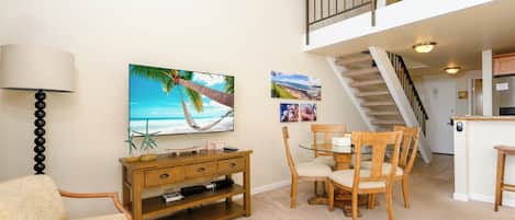 A living room with a TV, wooden furniture, a dining table, and a staircase leading to an upper level.