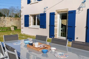Een glazen tafel met stoelen, een dienblad met kopjes en een schaal, en uitzicht op een gebouw met blauwe luiken en een stenen muur.