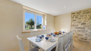 A dining area with a white table set for six, surrounded by white chairs, with a stone wall and a window overlooking a garden. - Ty Bidou, charming house near the sea (Kerlouan)