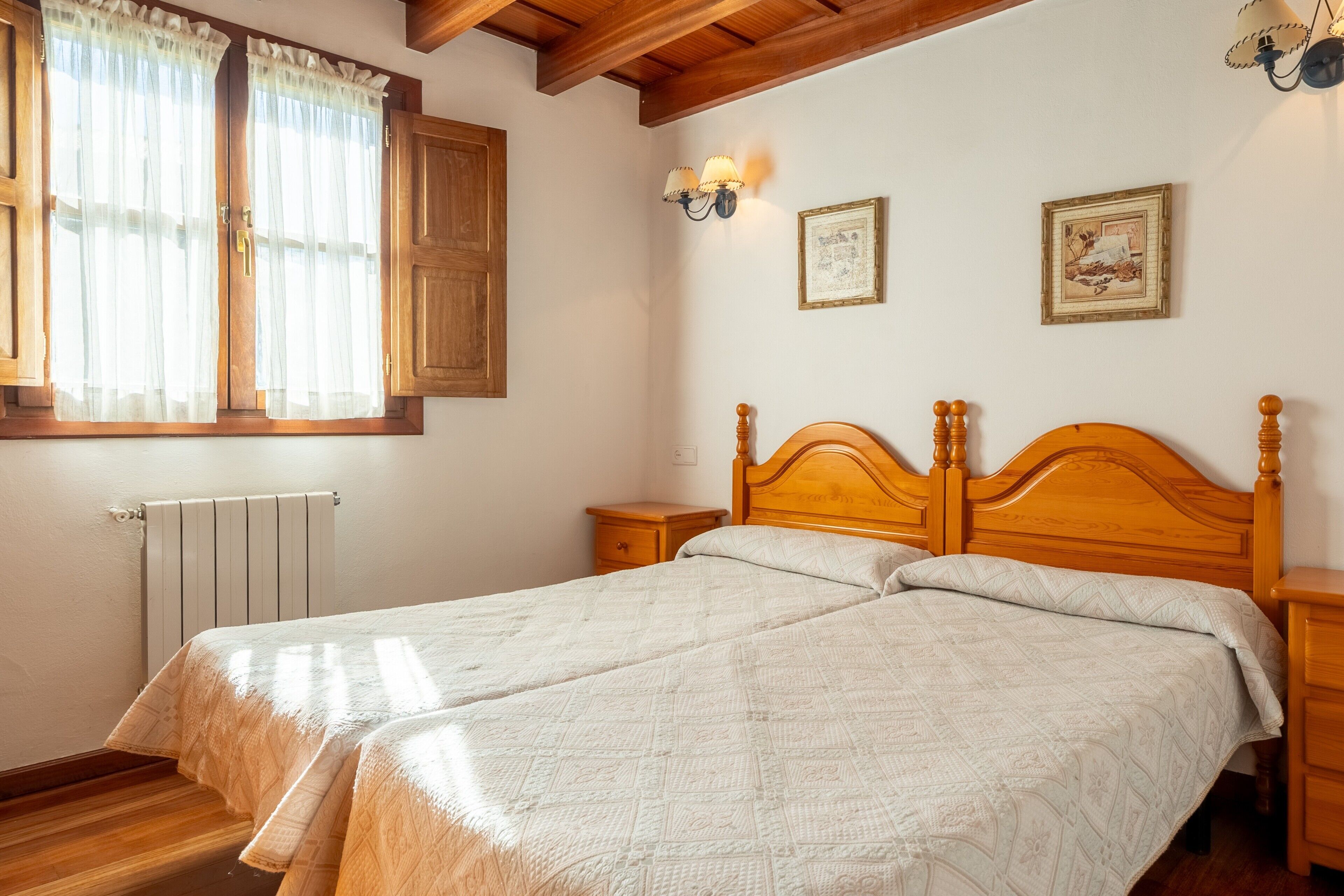 Un lit double avec une tête de lit en bois, une table de chevet, une commode, une fenêtre avec des rideaux et des appliques murales.