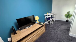 Una habitación con escritorio de madera, televisor de pantalla plana, lámpara y una pequeña mesa con sillas.