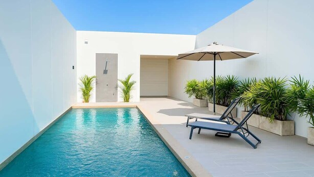 Un área de piscina moderna con una tumbona y una sombrilla, rodeada de plantas en macetas.