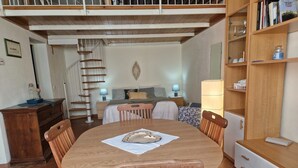 Een eetgedeelte met een houten tafel en stoelen, een spiltrap, een bed met hoofdeinde en een boekenkast.
