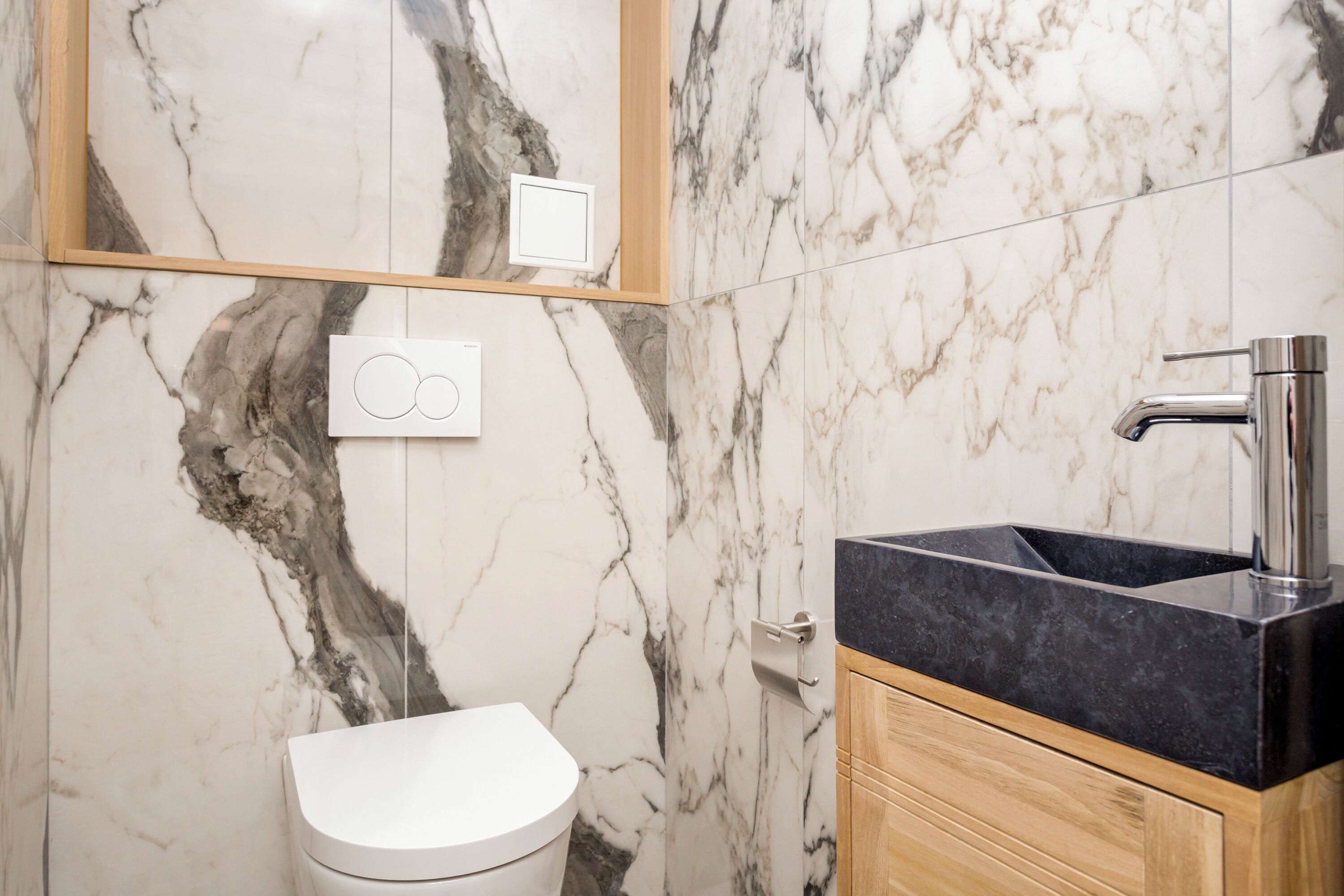 Ein Badezimmer mit Marmorwand, einer weißen Toilette, einem schwarzen Waschbecken und Holzschränken.