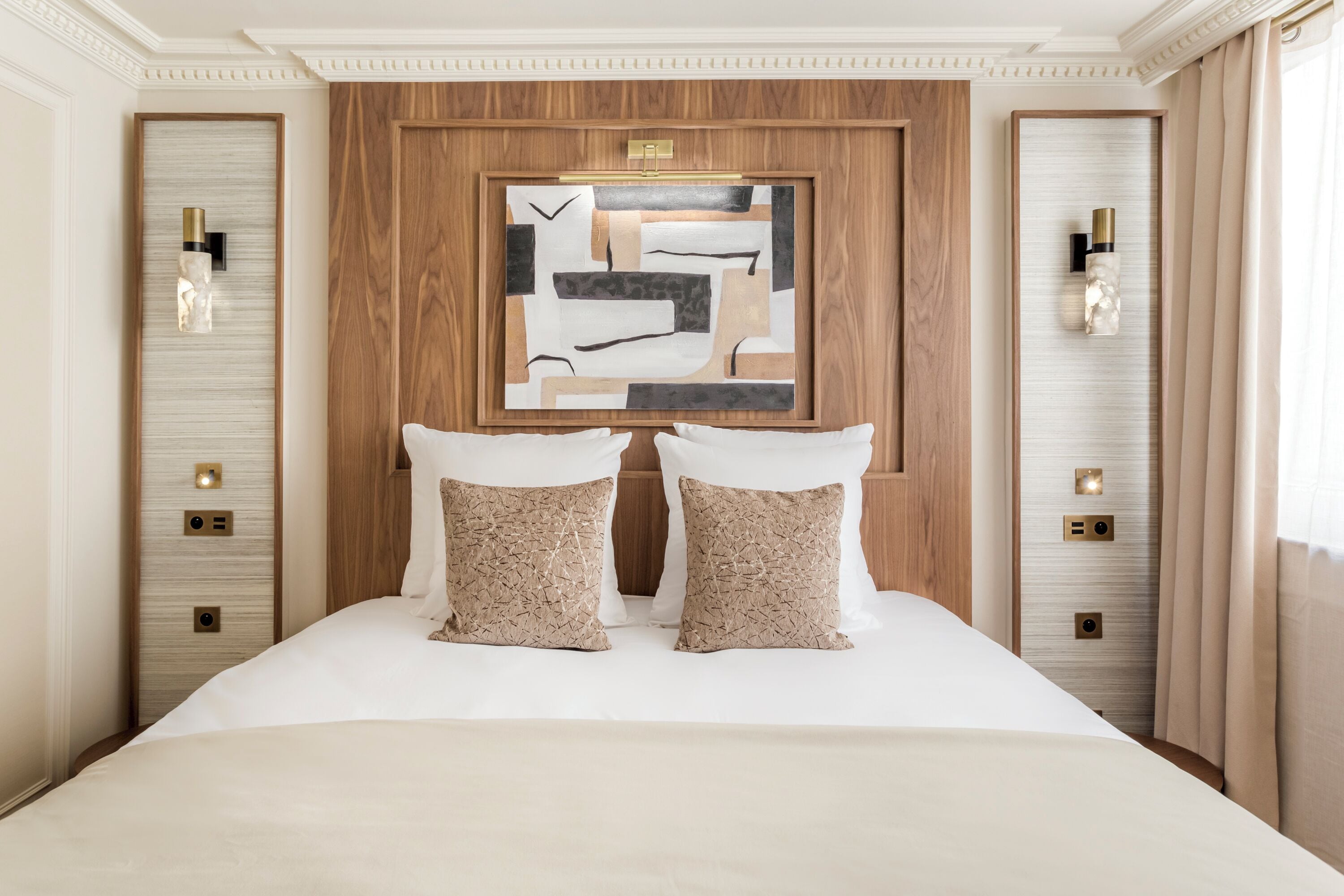 Quarto de hotel com uma cama grande, duas almofadas decorativas, uma obra de arte abstrata emoldurada acima da cama e luminárias fixadas na parede.