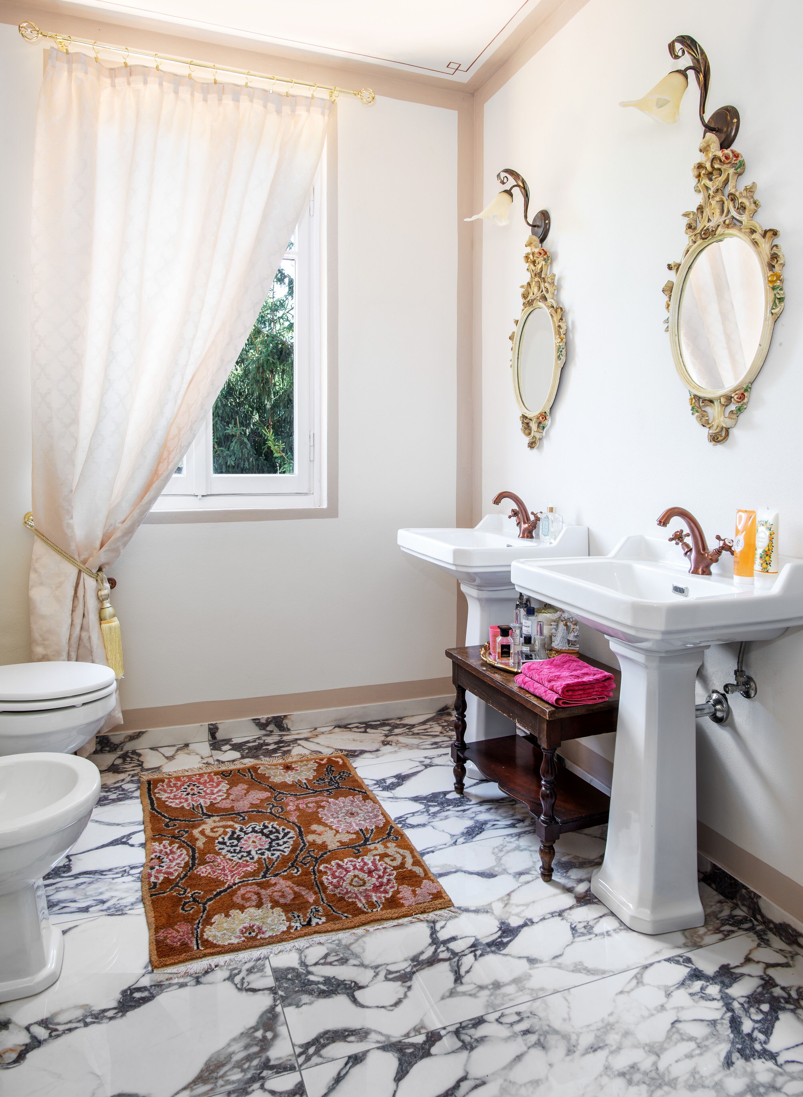 A bathroom with a toilet, two sinks, a mirror, a window with a curtain, and a patterned rug.