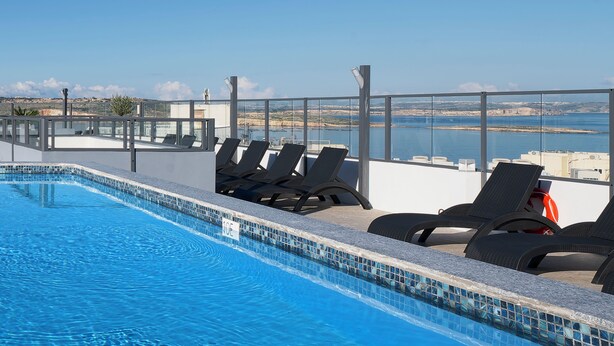 A rooftop pool with clear blue water, surrounded by lounge chairs and a glass balustrade offering a view of the sea.