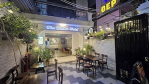 An outdoor seating area with tables and chairs, a neon sign for 'The Dahlia Hotel', and a black gate to the right.