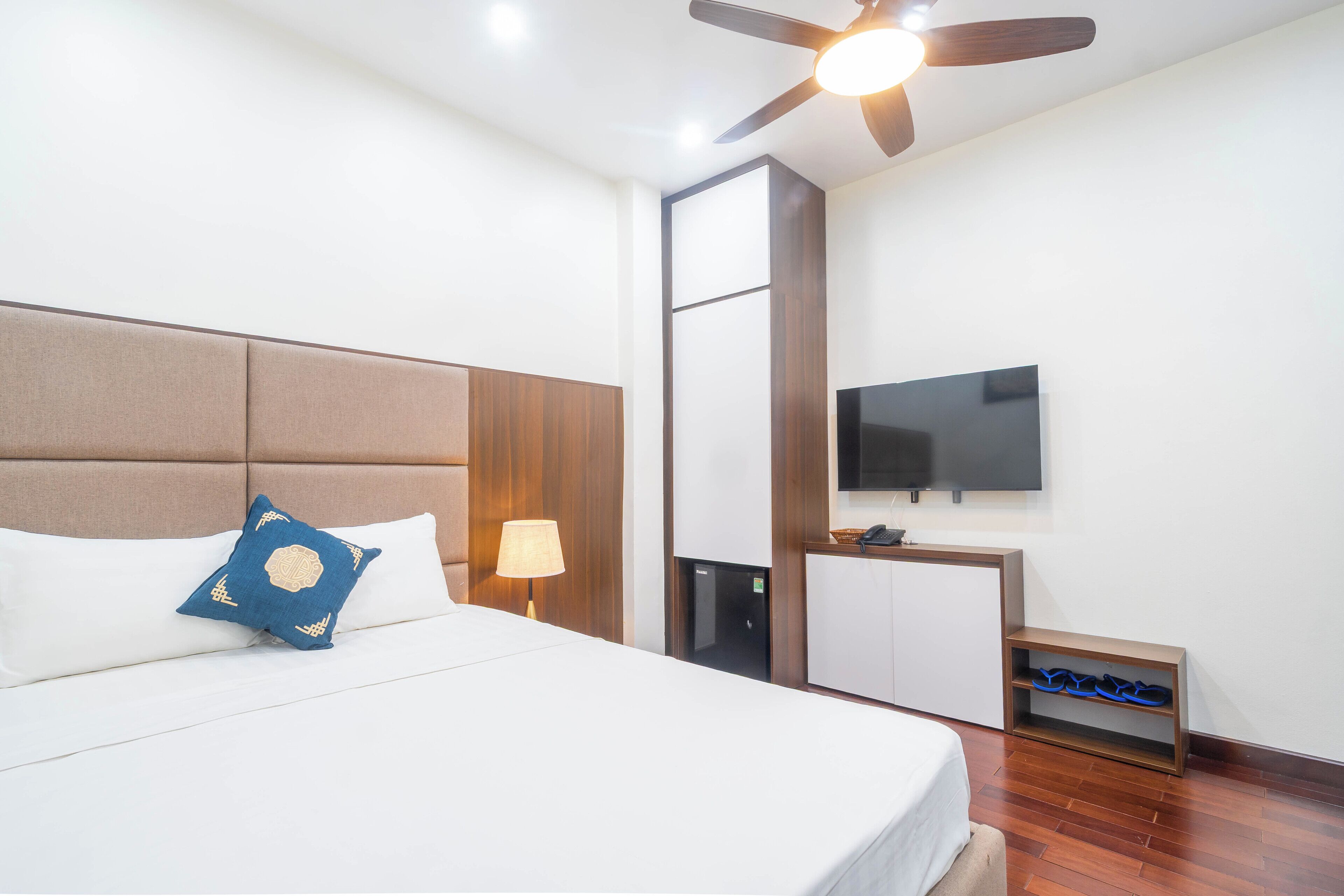 A hotel room with a large bed, a ceiling fan, a TV, a minibar, and a shoe rack.