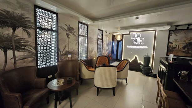 A modern hotel lobby with a seating area, palm tree murals, and a sign for 'The Dahlia Hanoi Hotel & Apartment'.