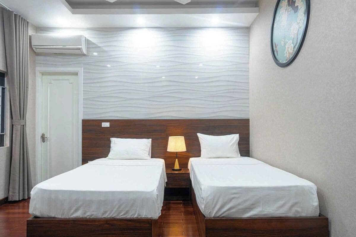 Ein Hotelzimmer mit zwei Einzelbetten, einem Nachttisch mit Lampe, einer Wandklimaanlage und einer Tür mit Vorhang.