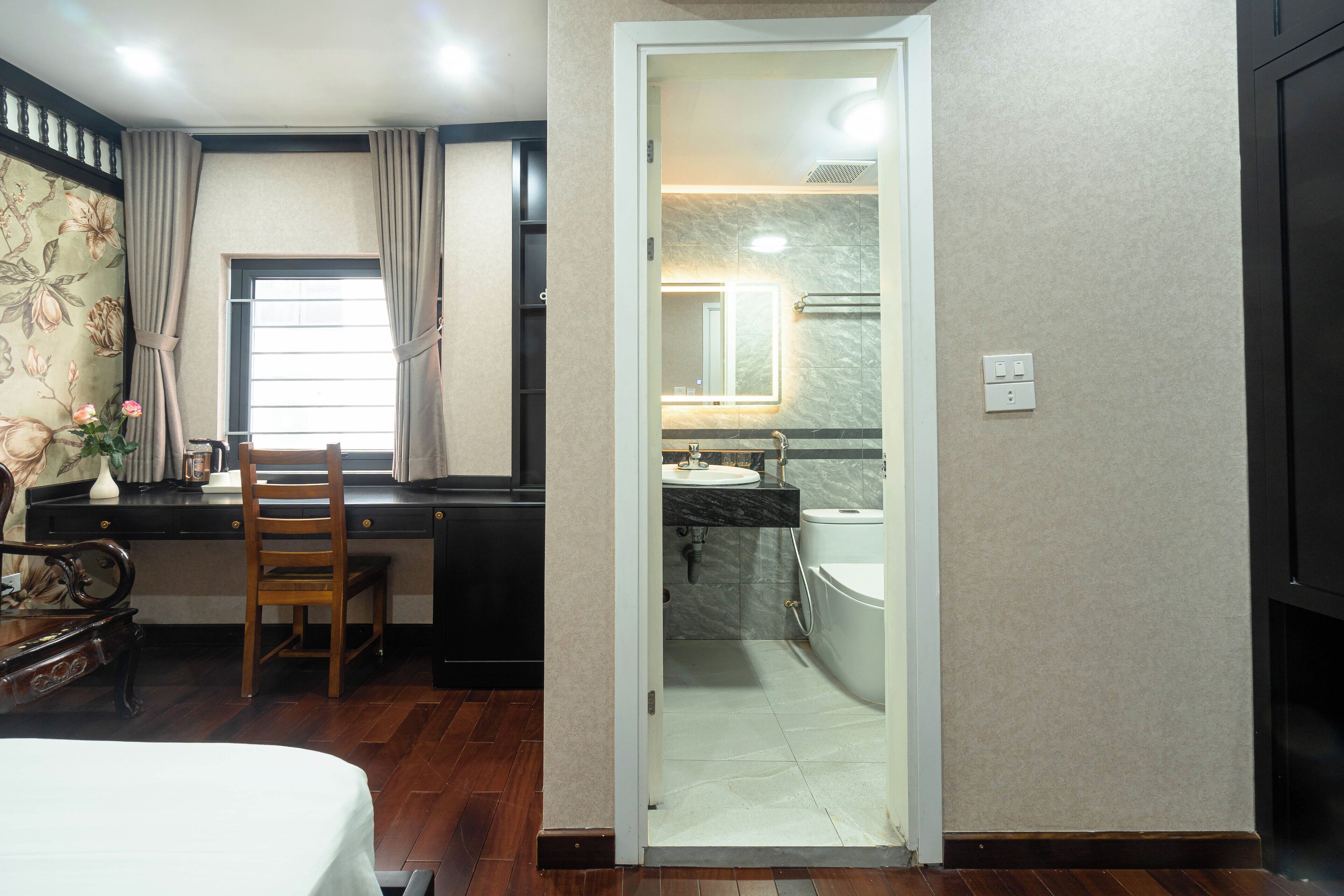Une chambre d’hôtel avec un bureau en bois, une chaise, un lit, une fenêtre avec des rideaux et une salle de bain équipée d’un lavabo et d’un miroir.