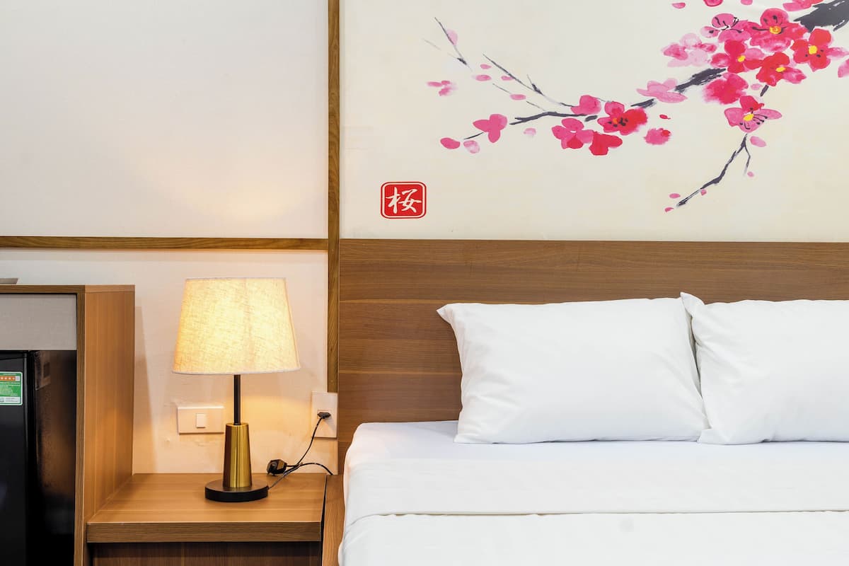 Ein Hotelzimmer mit einem Bett, weißen Kissen, einem Nachttisch mit Lampe und einem an der Wand hängenden Gemälde von Kirschblüten.